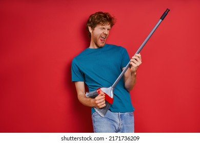 Playful Young Redhead Man Using Broom Like Guitar While Against Red Background