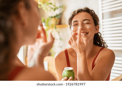 Playful young woman applying cream on nose. Girl holding green lotion jar applying moisturizer on nose. Beautiful woman taking care of skin by applying moisturizer every day in the morning.