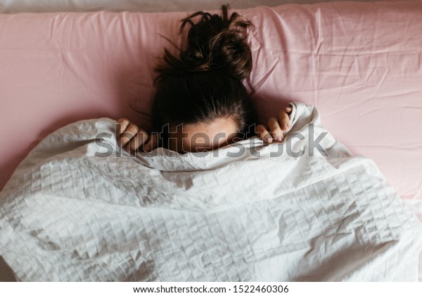 Playful young\
woman hiding face under blanket while lying in cozy bed, pretty\
curious girl feeling shy peeking from duvet, covering with white\
sheet, head shot close up. Top\
view