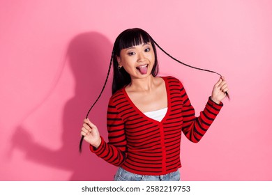 Playful young woman pulling braided hair and sticking out her tongue against a bright pink background