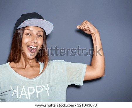 Playful young woman pumping her muscles flexing her arm to show off her biceps with a laughing smile, over grey