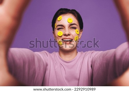 Playful young woman taking a selfie with smiley stickers on her face. Happy woman smiling at the camera while taking a picture of herself. Cheerful young woman having fun against a purple background.