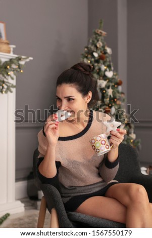 playful young woman in a transparent sweater with ginger cookies and a cup of coffee sits by the New Year tree