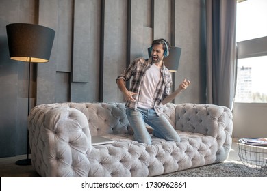 Playing An Air Guitar. Bearded Young Man In Headphones In Jeans Kneeling On The Couch Playing An Air Guitar, Closing His Eyes.