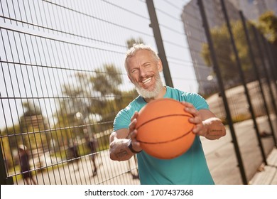 Playing Basketball. Cheerful Middle Aged Man In Sportswear Holding Basketball Ball And Smiling At Camera While Standing At Outdoor Basketball Court