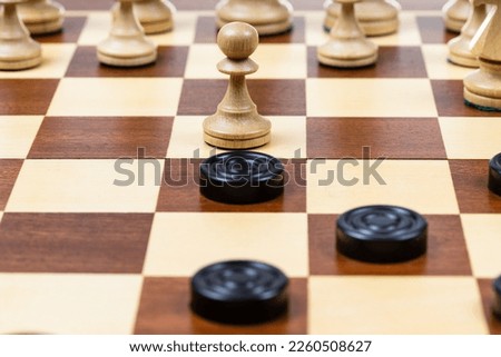 playing by different rules on the same board - black checkers and white chess figures on wooden chessboard, pawn and checkers piece in center of playfield closeup (focus on the pawn)