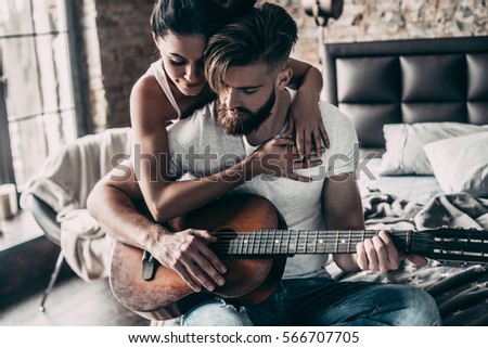 Playing by heart. Handsome young bearded man sitting in bed and playing guitar while attractive woman embracing him 