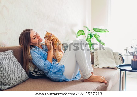 Playing with cat at home. Young woman sitting and relaxing on couch in living room and hugging, playing with pet.