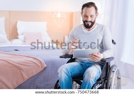 Playing computer games. Content cute bearded disabled man smiling and holding a tablet while sitting in a wheelchair