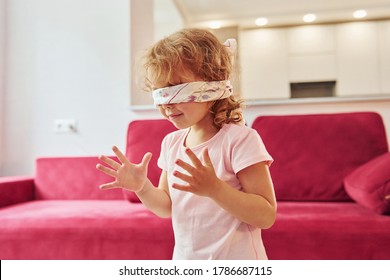 Playing game with eyes blindfolded. Cute little girl in casual clothes is indoors at home at daytime.