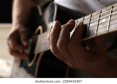 Playing the guitar. Strumming acoustic guitar. Musician plays music. Man fingers holding mediator. Man hand playing guitar neck in dark room. Unrecognizable person rehearsing, fretboard close-up.
