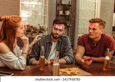 Playing hedbanz game together. Attractive joyous active good-looking vigorous young-adult radiant friends enjoying time at the bar while playing a hedbanz game together