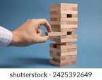 Playing Jenga. Man removing wooden block from tower on blue background, closeup