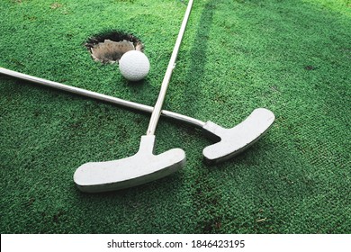 Playing Mini Golf On Artificial Green Grass Outdoors. Setting Goals And Winning Concept For Family Or Couple. Fun Vacation Entertainment. Two Sticks And Ball Near Target Hole On Golf Course Outside.