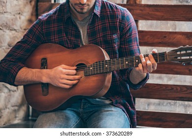Playing perfect riff. Close-up part of young man playing guitar while sitting at windowsill 
