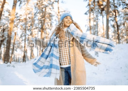 Playing with snow. Beautiful woman in a hat and scarf playing with snow, having fun in a snowy winter forest. Cheerful woman enjoying sunny frosty weather.