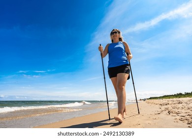 Playing sports on beach in summer. Portrait of young modern woman with long blonde hair in blue top and black shorts and headphones, sunglasses practicing Nordic walking on sandy beach on sunny day.  - Powered by Shutterstock