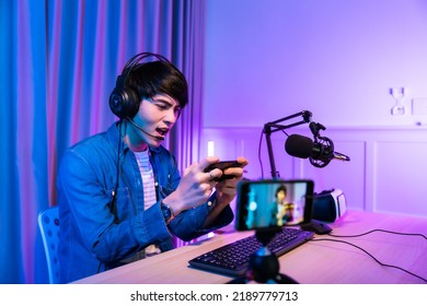 Playing Video Games On Smartphone. Young Asian Handsome Man Sitting On Chair Holding Cellphone In His Hand. Exited Streamer Online Mobile Game In Neon Room .Esport Streaming Game Online.