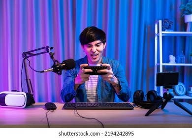 Playing Video Games On Smartphone. Young Asian Handsome Man Sitting On Chair Holding Cellphone In His Hand. Exited Streamer Online Mobile Game In Neon Room .Esport Streaming Game Online.