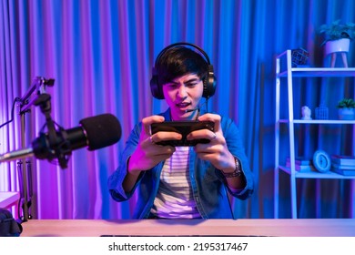 Playing Video Games On Smartphone. Young Asian Handsome Man Sitting On Chair Holding Cellphone In His Hand. Exited Streamer Online Mobile Game In Neon Room .Esport Streaming Game Online.