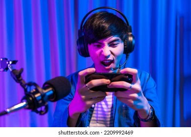 Playing Video Games On Smartphone. Young Asian Handsome Man Sitting On Chair Holding Cellphone In His Hand. Exited Streamer Wearing Headset In Online Mobile Game In Neon Room .Esport Streaming Game.