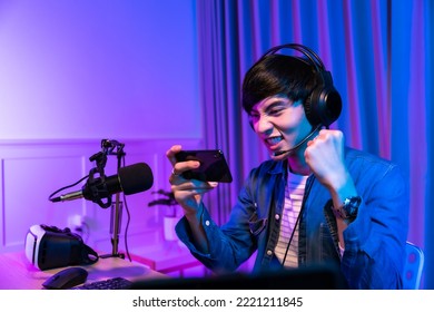 Playing video games on smartphone. Young asian handsome man sitting on chair holding cellphone in his hand. Exited streamer wearing headset in online mobile game in neon room . - Powered by Shutterstock