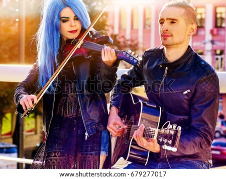 Playing viola woman and man perform music on violin and guitar in city outdoor. Girl with blue hairstyle and eyebrows performing jazz on urban street. Couple of musicians in love makes living.