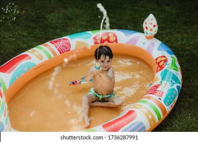 Playing With Water Gun In An Inflatable Swimming Pool At Home In A Garden, Family Fun During Lockdown And Quarantine