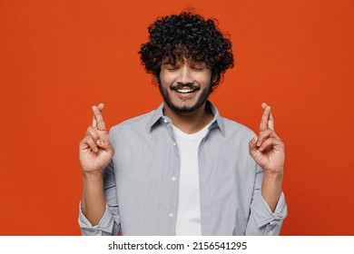 Pleading jubilant young bearded Indian man 20s years old wears blue shirt waiting for special moment keeping fingers crossed making wish eyes closed isolated on plain orange background studio portrait