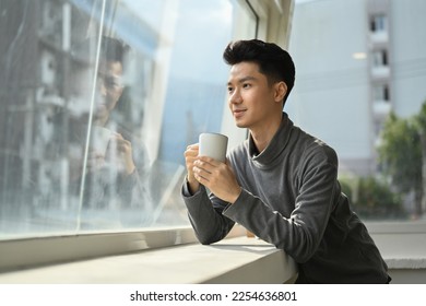Pleasant asian man enjoying his morning coffee while standing near office window overlooking cityscape - Shutterstock ID 2254636801