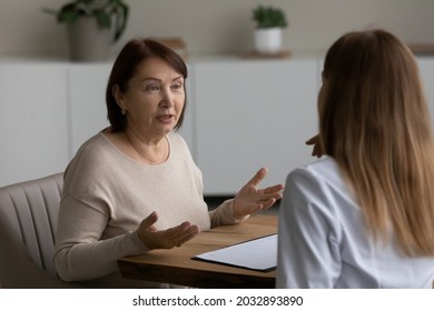 Pleasant Beautiful Middle Aged Older Retired Woman Telling Health Complaints Or Discussing Healthcare Issues With Young Female General Practitioner Doctor Physician At Checkup Meeting In Clinic.