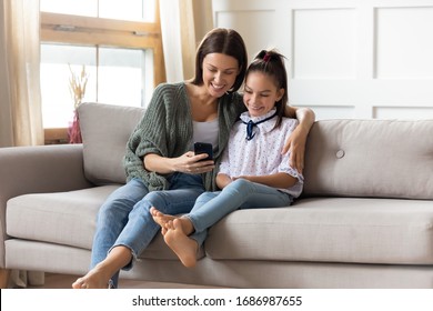 Pleasant beautiful young woman embracing adorable kid girl, showing educational application on smartphone. Smiling mother shopping online with happy schoolgirl daughter via cellphone, sitting on sofa.