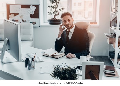 Pleasant Business Talk. Handsome Young Man In Full Suit Talking On The Phone And Looking Away While Sitting At The Office Desk 