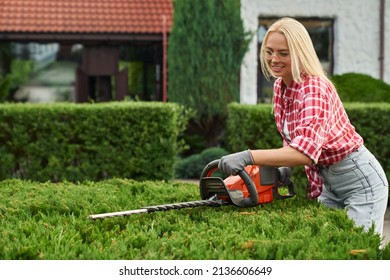 Pleasant Caucasian Woman In Casual Clothes, Safety Glasses And Gloves Pruning Bushes With Electric Trimmer During Summer Time. Gardening Concept.