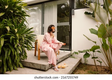 Pleasant Caucasian Young Brunette Woman Sits On Armchair In Backyard Summer. Girl Wears Sunglasses, Pink Shirt And Pants. Leisure Lifestyle And Beauty Concept.