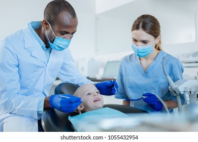 Pleasant Collaboration. Two Young Skilled Dentists Conducting A Dental Treatment With Preliminary Oral Cavity Checkup While The Girl Smiling