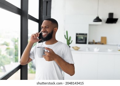 Pleasant Conversation. Portrait of smiling African American man talking on mobile phone in the morning standing near window holding cup of hot coffee, making call with friends or family - Powered by Shutterstock