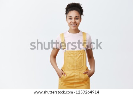 Pleasant cute female dark-skinned employee helping customers find right item to purchase. Joyful friendly-looking girl in trendy yellow overalls, holding hands in pockets and smiling at camera