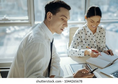 Pleasant distraction. Charming young man sitting at the table next to his female colleague and checking his phone while the woman making notes