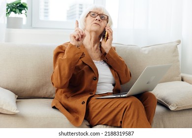 A Pleasant, Emotional Old Lady Is Sitting On A Sofa In A Beautiful Apartment In A Stylish Brown Suit Talking On The Phone With A Laptop On Her Lap, Holding Her Index Finger Up