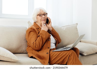 A Pleasant, Emotional Old Lady Is Sitting On A Sofa In A Beautiful Apartment In A Stylish Brown Suit Talking On The Phone With A Laptop On Her Lap, Holding Her Index Finger Up