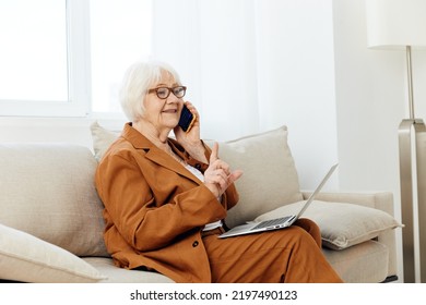 A Pleasant, Emotional Old Lady Is Sitting On A Sofa In A Beautiful Apartment In A Stylish Brown Suit Talking On The Phone With A Laptop On Her Lap, Holding Her Index Finger Up