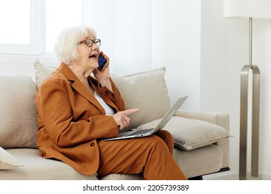 A Pleasant, Emotional Old Lady Is Sitting On A Sofa In A Beautiful Apartment In A Stylish Brown Suit Talking On The Phone With A Laptop On Her Lap, Holding Her Index Finger Up