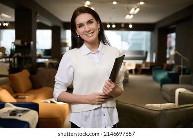 Pleasant European Pretty Woman, Retail Assistant And Sales Rep Holding Catalog With Upholstered Furniture, Standing In The Exposition Center And Smiling With Cheerful Toothy Smile Looking At Camera