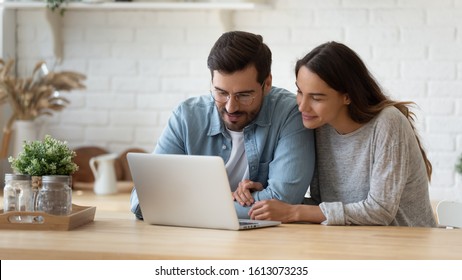 Pleasant family couple sitting at big wooden table in modern kitchen, looking at laptop screen. Happy young mixed race married spouse web surfing, making purchases online or booking flight tickets. - Powered by Shutterstock
