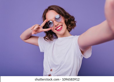 Pleasant girl with tattoo making selfie in studio and laughing. Good-looking young woman with brown wavy hair taking picture of herself on bright purple background. - Shutterstock ID 1096583912