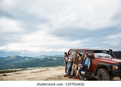 산 언덕에서 전망을 즐기면서 자동차 근처에 서 있는 즐거운 젊은 친구들 스톡 사진