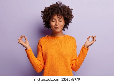 Pleasant Looking Calm Woman Meditates Indoor, Holds Hands In Mudra Gesture, Has Charming Smile, Closed Eyes, Wears Orange Clothes, Models Over Purple Background. Hand Gesture. Meditation Concept