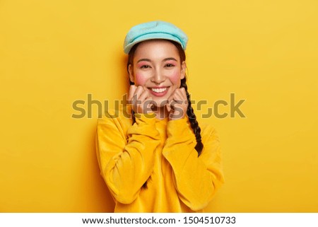 Pleasant looking cheerful Asian girl keeps both hands under chin, has pinup makeup, wears blue stylish cap, velvet yellow hoody, isolated, expresses positive emotions, has charming smile, two pigtails