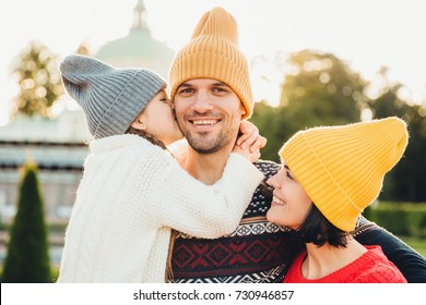 Pleasant Looking Little Girl Being Thankful To Her Father For Buying New Bicycle, Kisses Him In Cheek. Young Family Walk Together Outdoor, Spend Spare Time In Park, Wear Knitted Fashionable Hats